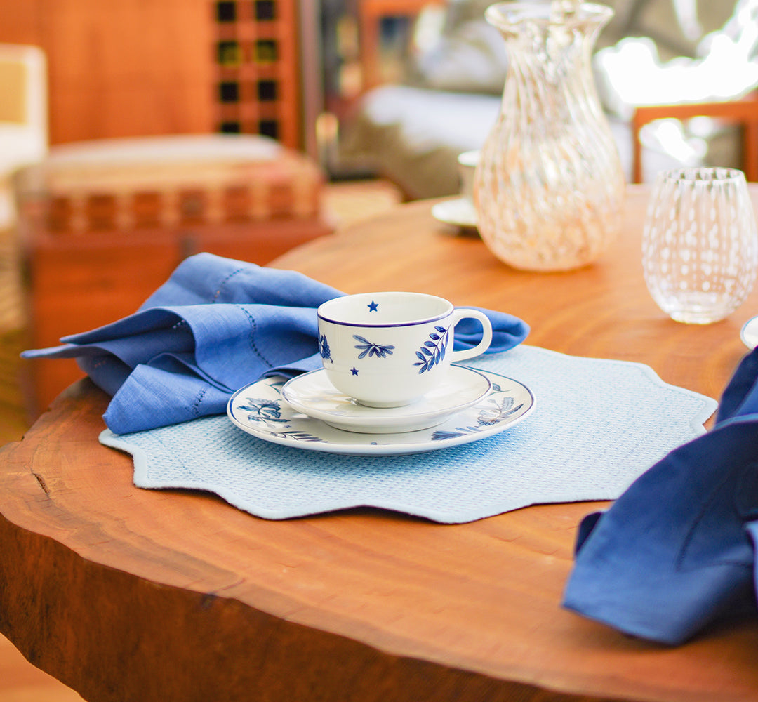 Jogo americano rafia redondo azul claro com xícara de porcelana floral e guardanapo azul em mesa de madeira com copos de cristal ao fundo