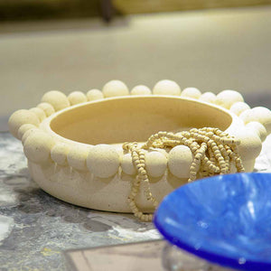 Centro de mesa redondo de cerâmica na cor marfim (natural), apresentando uma textura externa rica composta por diversas esferas em relevo. A peça possui tamanho de 35 cm de largura e 10 cm de altura, com design atemporal e acabamento rústico que destaca as variações únicas de cada peça. Como deve ser usado: ideal para compor a decoração de mesas de jantar ou centros de sala, sendo perfeito para acomodar frutas, esferas decorativas ou ser usado como uma peça de destaque chic e com personalidade.
