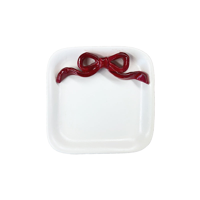 Pratinho redondo de cerâmica branca artesanal com detalhe decorativo de laço pintado em vermelho na borda. A peça possui 17 cm de diâmetro e 2 cm de altura, apresentando textura suave e acabamento manual exclusivo da coleção Lolahome.